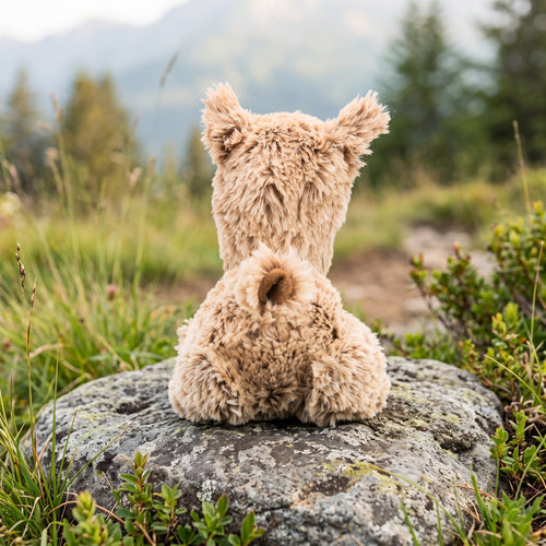 Vue de dos d'une peluche de bébé lama moelleuse installée sur un rocher dans un paysage alpin verdoyant, mettant en valeur la texture de sa fourrure et sa petite queue.